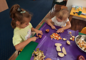 Dzieci siedzą przy stole i kroją owoce na sałatkę.