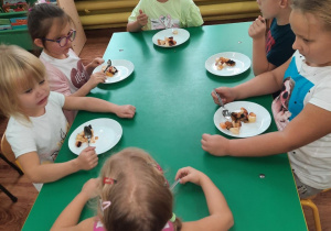 Dzieci siedzą przy stole i zjadają własnoręcznie przygotowaną sałatkę.