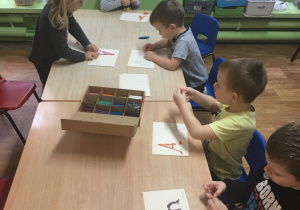 Dzieci siedzą przy stole i wylepiają literę "A, a".