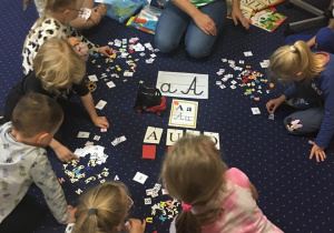 Dzieci siedzą na dywanie i tworzą napisy z liter.