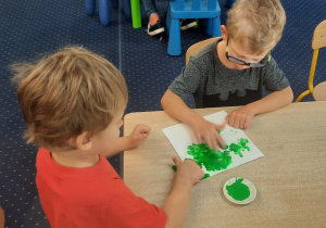 Chłopcy siedzą przy stole i stemplują palcami z wykorzystaniem farb kształt kwadratu.