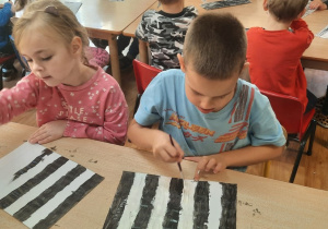 Dzieci siedzą przy stolikach i malują farbami pasy na kartonie.