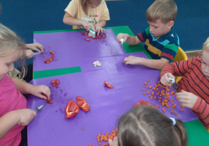 Dzieci siedzą przy stole i kroją warzywa.