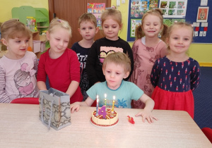 Chłopiec siedzi przy stole przed tortem, a pozostałe dzieci stoją za nim.