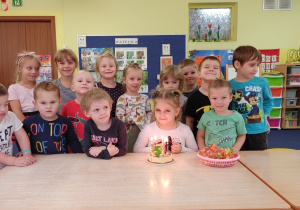 Dziewczynka siedzi przy stole przed tortem, a pozostałe dzieci stoją za nią.