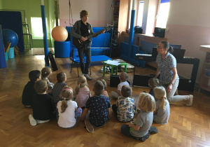 Dzieci siedzą na podłodze i słuchają ucznia grającego na gitarze elektrycznej.