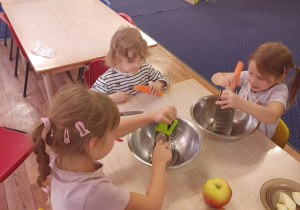 Dzieci siedzą przy stole i trą marchewkę i jabłko.