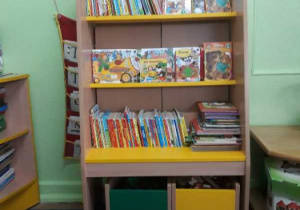 Kącik czytelniczy w którym stoi stojak wypełniony książeczkami dla dzieci.