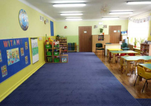 Widok na całą klasę od strony drzwi wejściowych. Po lewej tablica z napisem "WITAMY", po prawej stoliki dzieci wraz z krzesłami za którymi stoi biurko nauczyciela. W oddali drzwi do łazienki i schowka