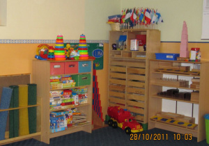 Kącik z pomocami i zabawkami. Pomoce montessoriańskie wraz ze stojakiem z dywanikami, duże auto i szafka z grami i puzzlami dla dzieci.