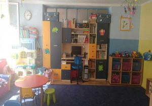 Na niebieskiej ścianie znajduje się zestaw mebli i szafka z zabawkami, pomocami dydaktycznymi, wnęka z biurkiem , na którym stoi komputer.na pierwszym planie stolik z krzesełkami.